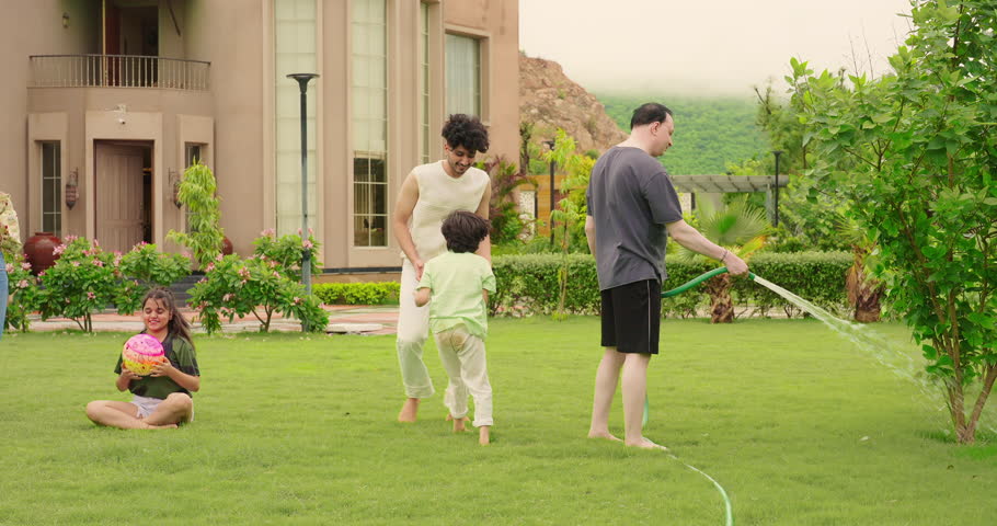 Indian happy father hold hose pipe spraying water on extended family group playing games in outdoor house backyard lawn having fun joy spend funny time enjoying summer vacation together outside park. 