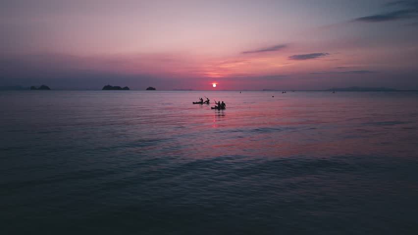 Tourists are enjoying peaceful kayaking experience in calm tropical sea during vibrant sunset, with silhouettes of islands adding to scenic beauty. Kayakers drifting through burning sunset glow