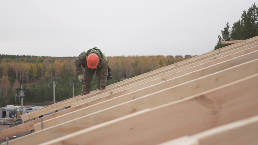 Builder hammers wooden roof frame under open sky surrounded by calm forest landscape. Natural backdrop highlights precise teamwork