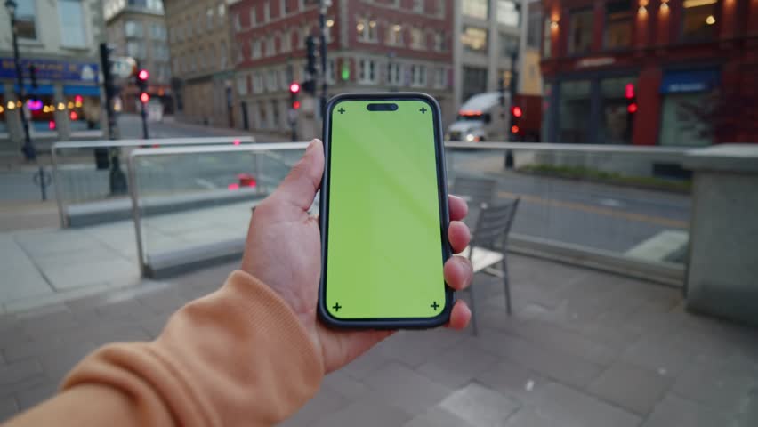A Bright City Day In Halifax, Canada, Where A Man Holds A Smartphone With Green Screen While Cars Pass And Tall Modern Buildings Line The Street.