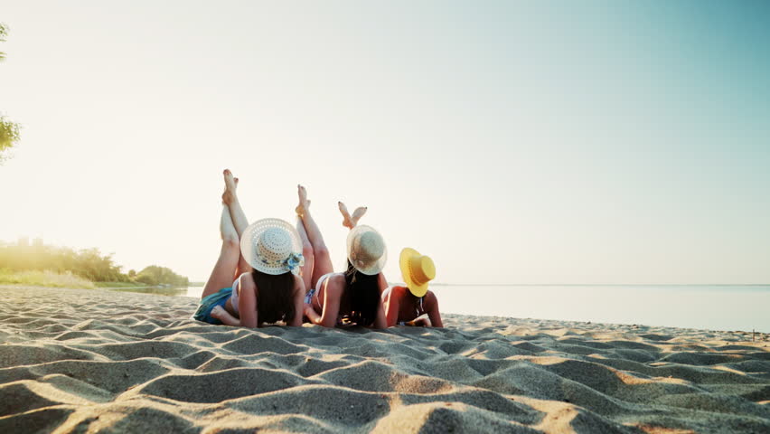 Three young beautiful girls are lying on a sandy beach and sunbathing in the evening sun, with their legs raised and crossed. The women are having fun by the sea, chatting, laughing and smiling