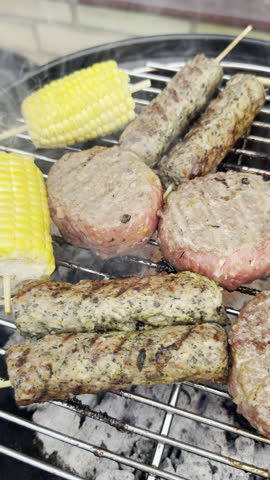 Vertical video - Closeup slow pan shot of burgers, kebabs, and cobs of sweetcorn cooking on a grate over hot flaming charcoal in an outdoor barbecue grill.