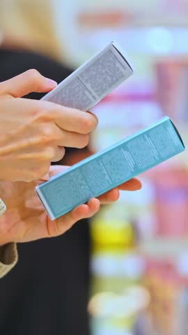 VERTICAL VIDEO Woman selecting a cosmetic serum for face or hair in a beauty store and reading ingredients, comparing products. Female customer holding skincare or haircare item. Conscious choice