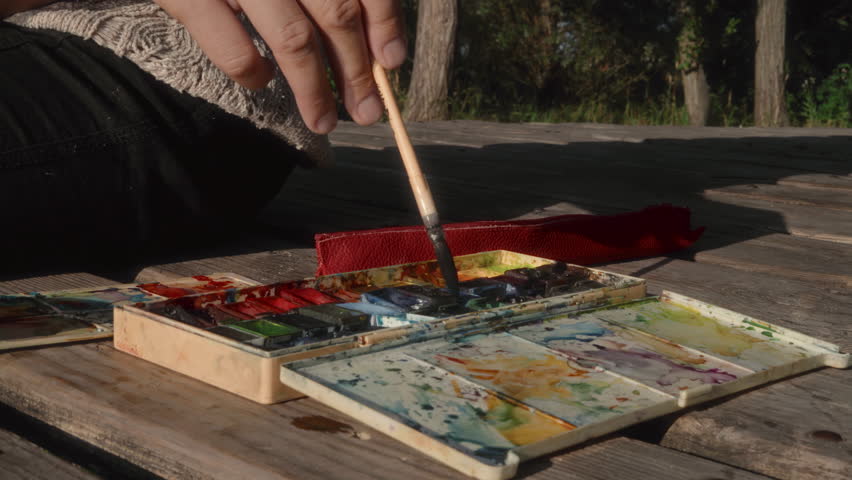 Close-up, female artist's hand picking up watercolor paint with brush on wooden pier