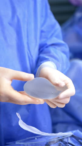 Gloved hands of the doctor wearing blue robe hold the breast implant. Medic turns the implant to the sides and takes it to the operating table. Vertical video.