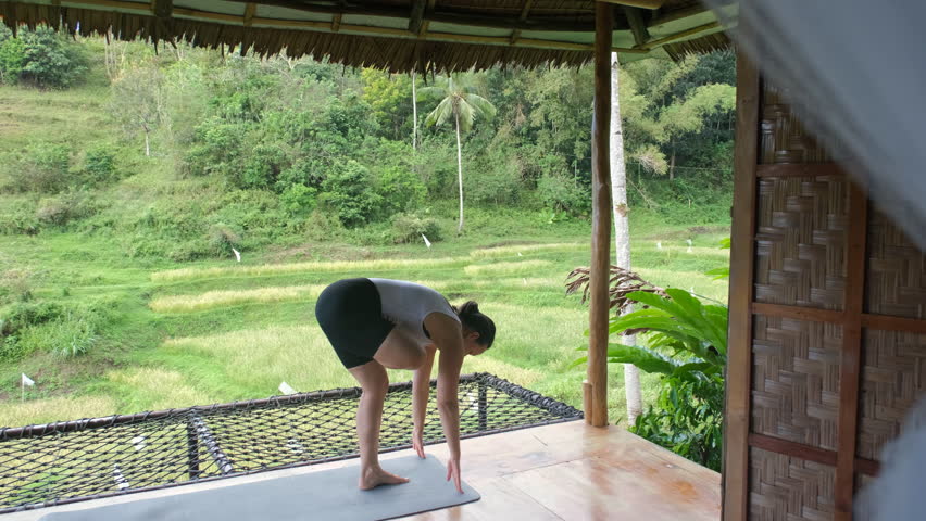 On a serene terrace of a bamboo hut, a young woman deeply engages in yoga while surrounded by breathtaking views of tropical greenery and rice plantations, slow motion