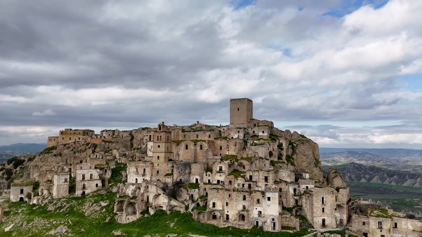 Aerial view of an ancient abandoned village of Craco, Italy with stone buildings and a tower on a hilltop, surrounded by lush greenery and distant mountains under a cloudy sky.