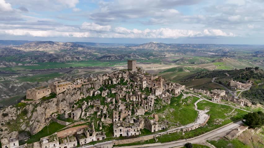 Aerial view of an ancient, abandoned hilltop village of Craco, Italy with crumbling stone buildings and a castle tower. The landscape is lush and green, under a cloudy sky.