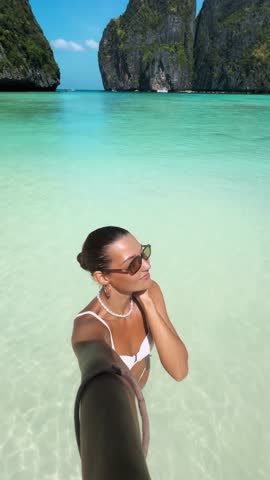 Stylish female blogger in white bikini films selfie video at tropical Maya Bay, smiling and blowing kiss to camera. Ideal for travel, influencer, lifestyle, and summer promo content.