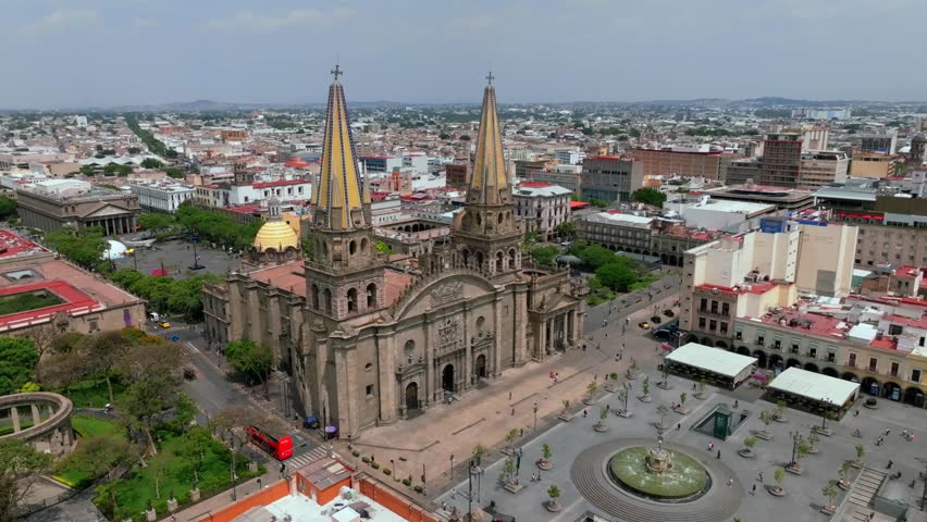 Wide aerial view of Catedral de Guadalajara facade with spires and plaza packed with tourists. Pan right reveaks Rotonda de los Jaliscienses Ilustres