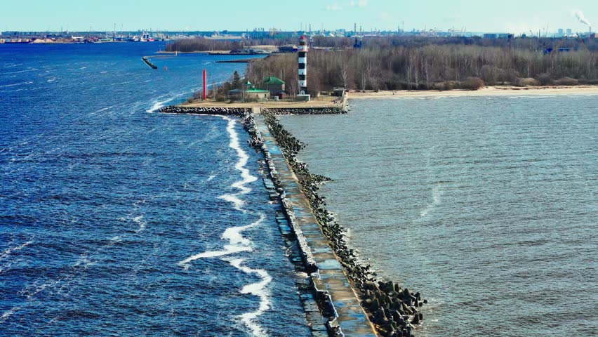 A bold black-and-white lighthouse stands at the end of a rugged stone pier, where vivid blue waves meet a tranquil sandy coastline edged by bare trees.