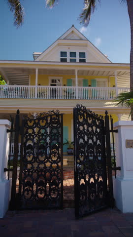 Vertical Screen: A breathtakingly beautiful yellow house featuring elaborate black gates, elegantly positioned amidst lush palm trees, set in a lively and bright tropical environment