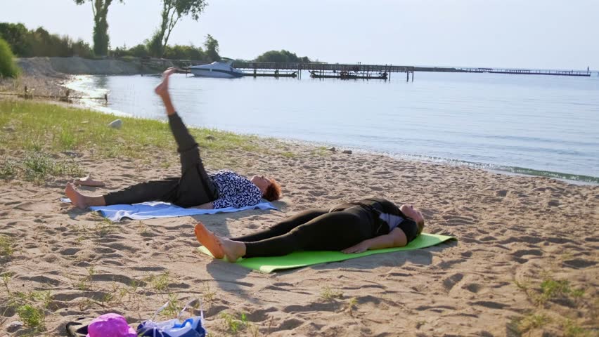 On a summer day by a scenic lake, a woman and her adult daughter slowly lift their legs, hold, then lower in a stretching routine. Moving outdoors eases tension and boosts flexibility.
