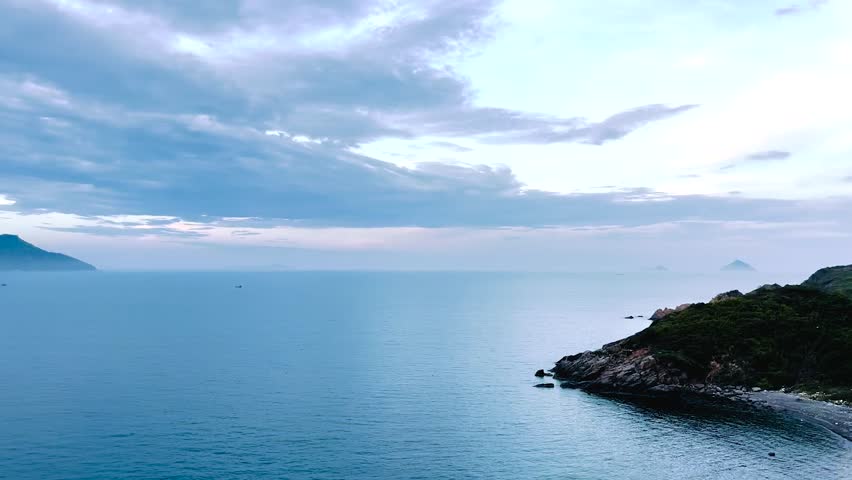 A serene aerial view of the ocean, with a calm coastline and distant hills in the background. The video captures the peaceful atmosphere of the sea under the cloudy sky, highlighting the natural beaut