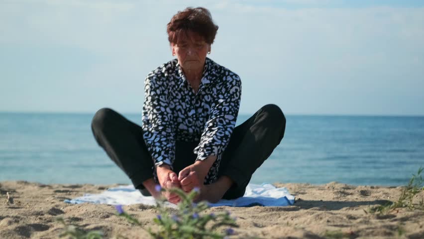 An senior woman sits in butterfly pose on warm sand at a summer lakeside beach. Holding her feet, she stretches hips and thighs, fully immersed in a relaxing outdoor yoga practice.