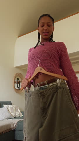 Vertical UGC footage of gen Z teenage African American girl vlogging on smartphone at home trying on streetwear cargo pants