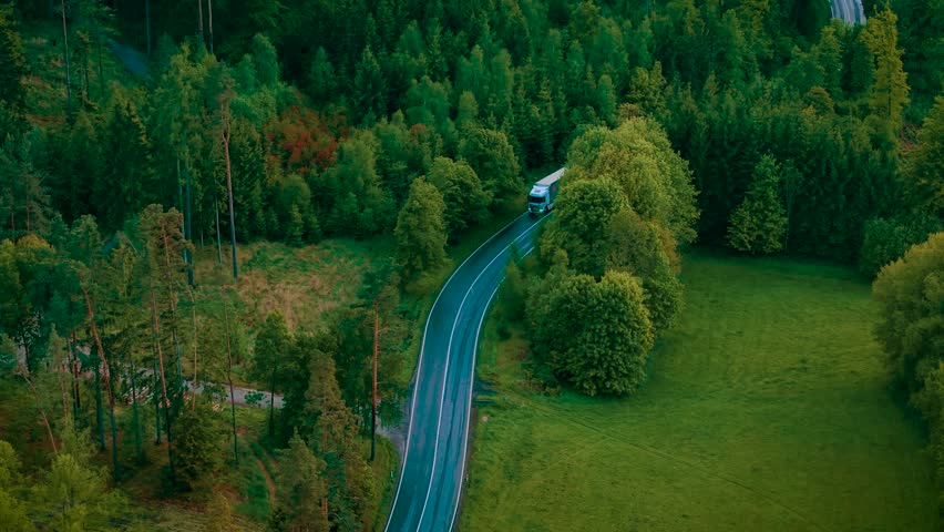 Truck driving on curvy forest road after rain. White semi truck moving along wet winding highway under overcast sky. Lorry traveling on slick asphalt route between trees and open greenery