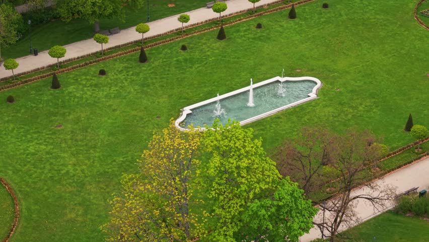 Top view of a decorative fountain with jets in a green manicured park garden surrounded by pathways, trees, and neatly trimmed bushes on a spring day