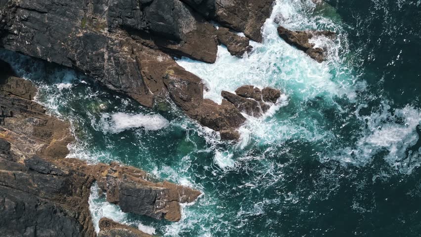 High birdseye aerial of turbulent waves crashing against Cornwall’s rocky shoreline with vivid green-blue water