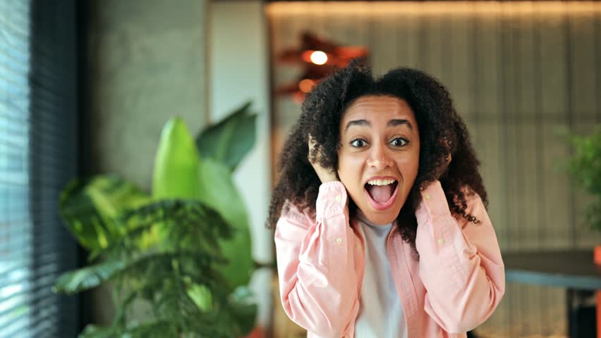 Excited African American female, dressed in a pink shirt and white t-shirt, girl shows extreme surprise and joy in a modern indoor office with green plants, expressing happiness.