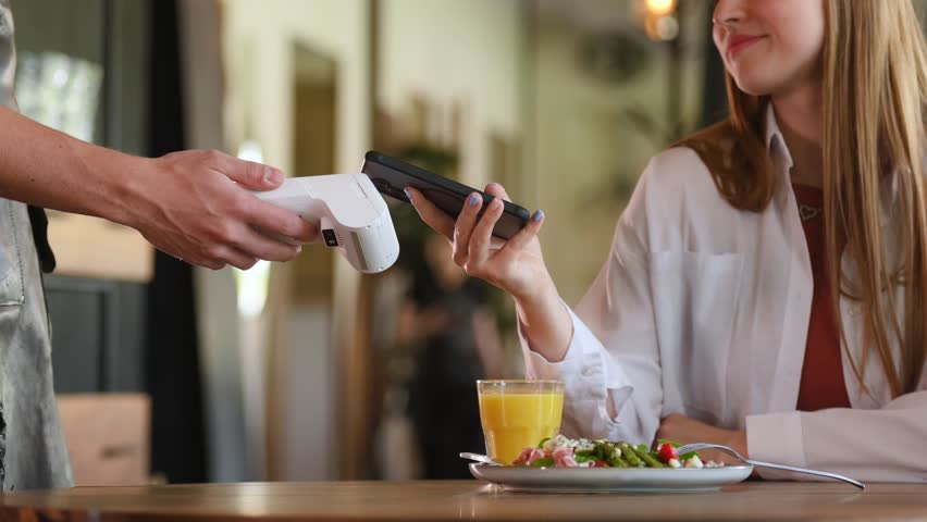A woman pays for her meal with a wireless payment by swiping her phone against a terminal in a cafe-restaurant.
