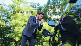 Stressed professional businessman in a suit gestures animatedly on a phone call outdoors. Beside black electric car charging in a green environment, upset specialist conveys frustration and urgency. - Powered by Shutterstock - Get 15% off with code: PIKWIZARD15