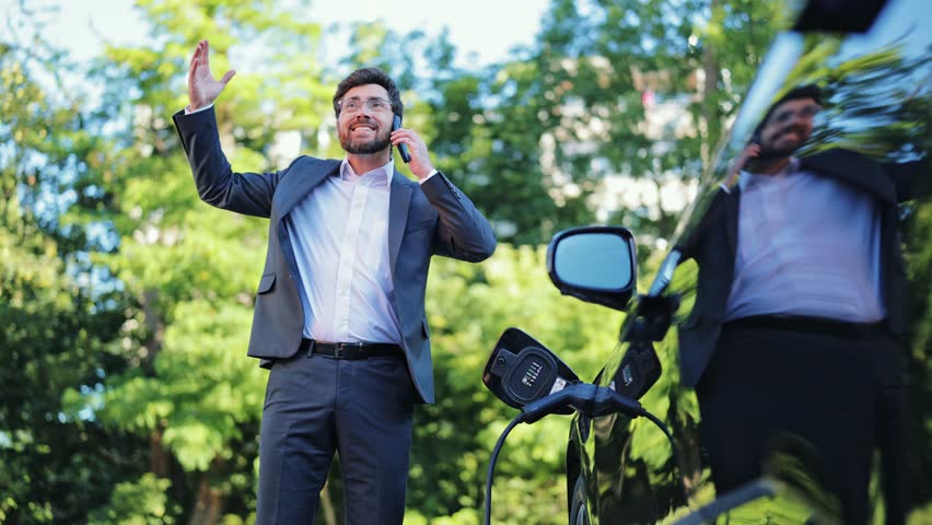 Stressed professional businessman, dressed in a grey suit, makes an urgent phone call outdoors beside a charging electric vehicle. Expresses frustration and concern during conversation.