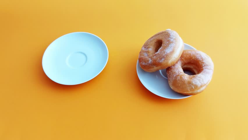 A hand reaches for a glass cup of milk placed on a saucer next to a plate of two glazed donuts on a vibrant orange background. Bright, minimalistic breakfast scene