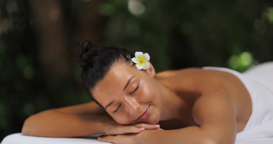 Caucasian woman lies on her stomach on a massage table with a plumeria in her hair, slowly opening her eyes and smiling in slow motion. Ideal for spa, wellness, and beauty lifestyle promos.