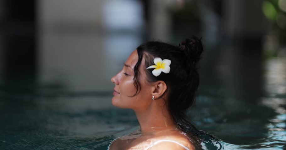 Caucasian woman with glowing skin and a plumeria flower in her hair gracefully rises from a pool in slow motion. Perfect for spa, wellness, luxury travel, and beauty lifestyle promotions.