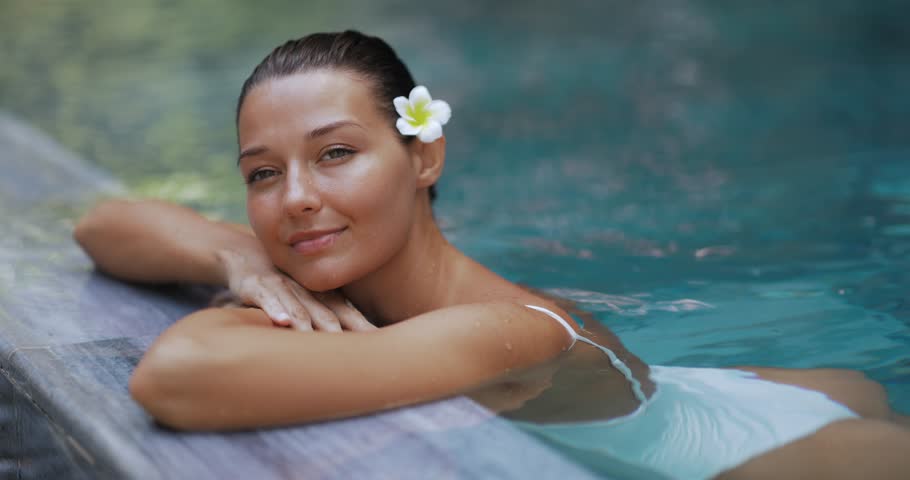 Slow motion concept of a tanned woman in a white swimsuit relaxing in a luxury tropical pool, with a plumeria flower in her hair. Ideal for beauty, travel, spa, and wellness promotions.