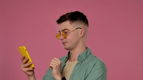 A man wearing yellow sunglasses scrolls through a feed on his smartphone against a pink background, creating a stylish and fashionable studio atmosphere. - Powered by Shutterstock - Get 15% off with code: PIKWIZARD15