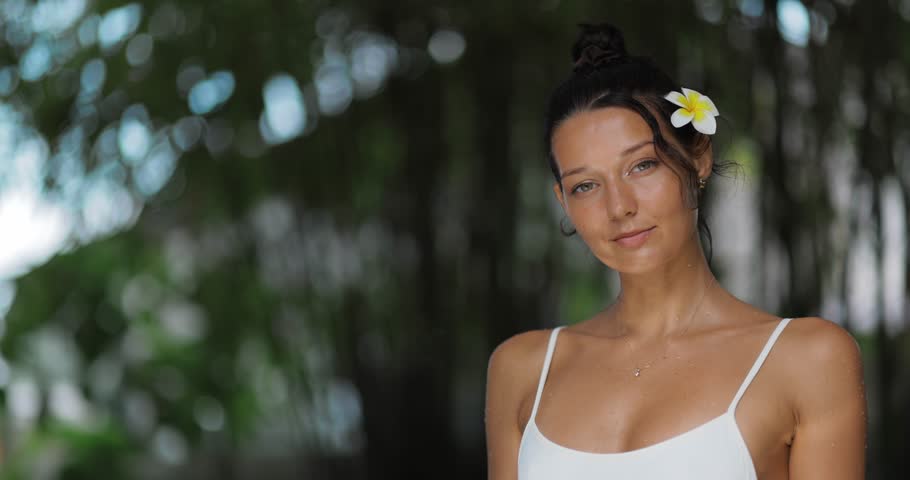 Trendy woman with glowing skin and tropical flower in hair poses by poolside in soft natural light. Ideal for beauty, spa, skincare, summer lifestyle or tropical vacation promo.