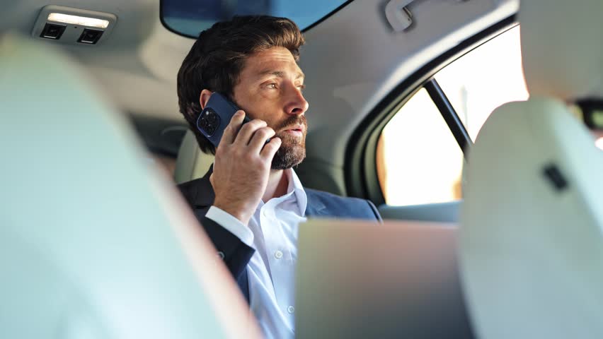 Professional businessman focuses during an important phone call while using a laptop in the luxurious back seat of an executive car. He appears busy and determined, managing business matters on the go