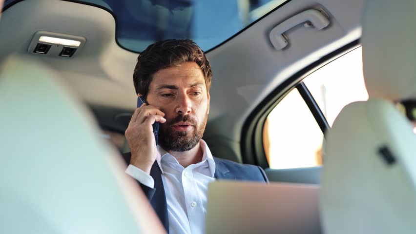 Professional businessman focuses during an important phone call while using a laptop in the luxurious back seat of an executive car. He appears busy and determined, managing business matters on the go