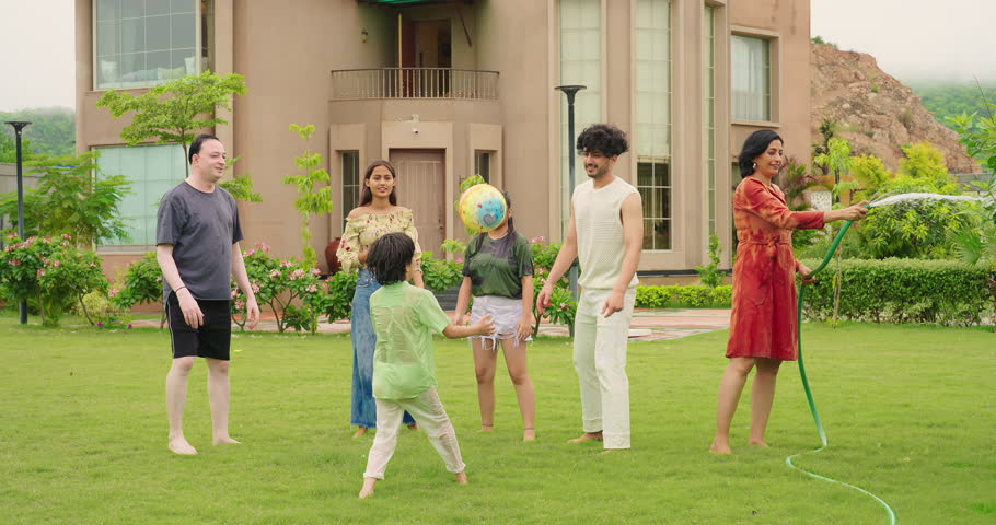 Indian happy mature mother hold hose pipe spraying water garden tree at outdoor house backyard. Extended family group play games passing ball enjoy together have fun joy spend funny time outside park