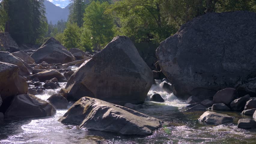 Footage of river water flowing between massive granite boulders on the Merced River in Yosemite National Park, California.