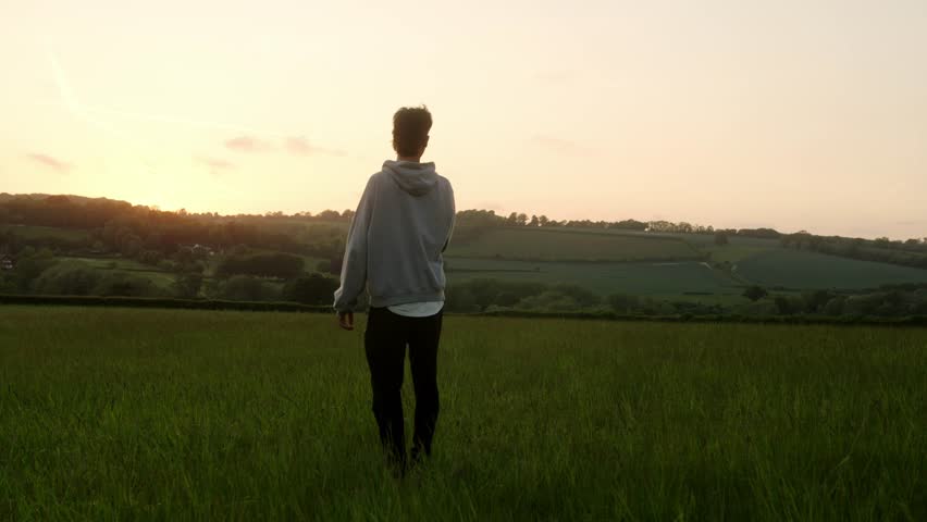 A man stands in a grassy field at sunset overlooking soft rolling hills
