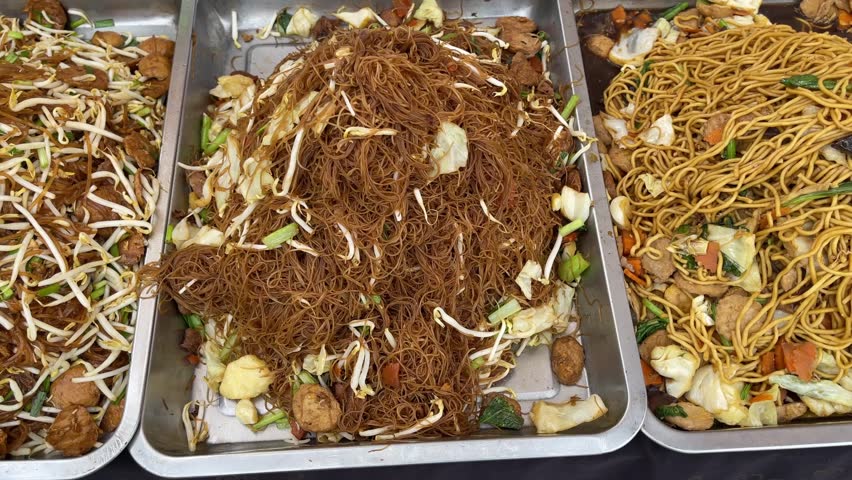 Assorted stir-fried Asian noodles with vegetables and tofu in stainless steel trays, including rice vermicelli, yellow noodles, and bean sprouts at Thai street food market