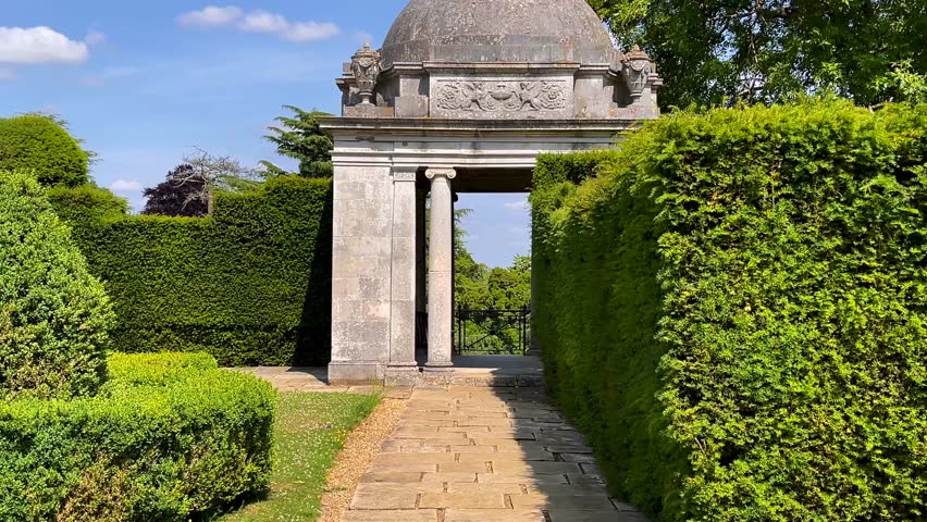 Garden pavilion framed by clipped green hedges and golden stone path beneath a blue English sky, traditional and timeless, slow motion