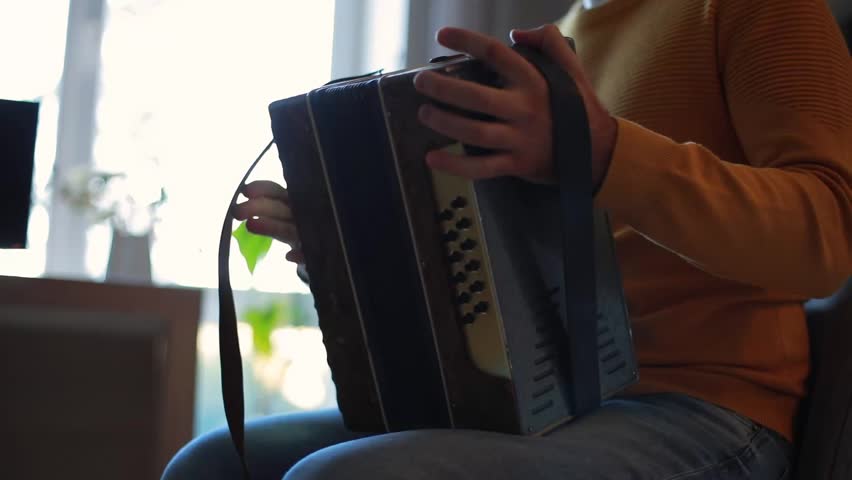 Man in mustard sweater playing accordion near window with natural light, close-up on hand and instrument, cozy creative atmosphere, warm home interior, music lifestyle.