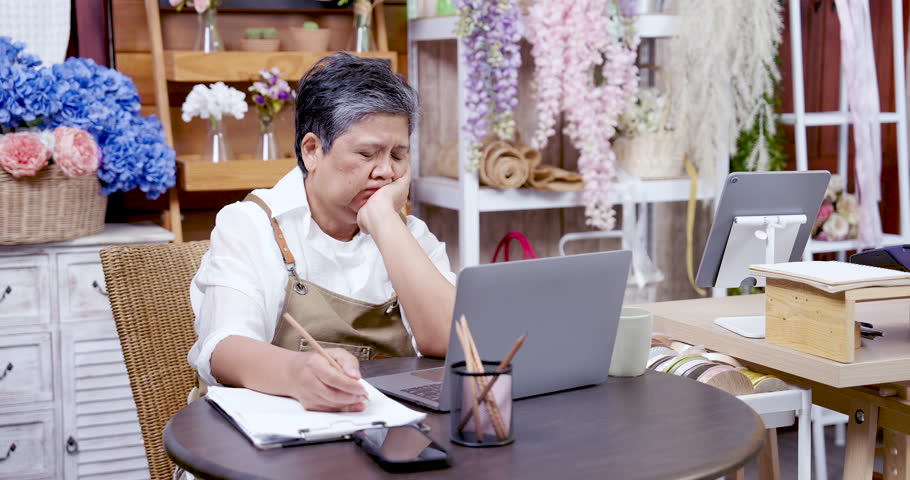 Asian elderly female florist sitting in floral shop looking at laptop with worried face while reviewing documents, expressing stress and pressure in running a small online flower business alone