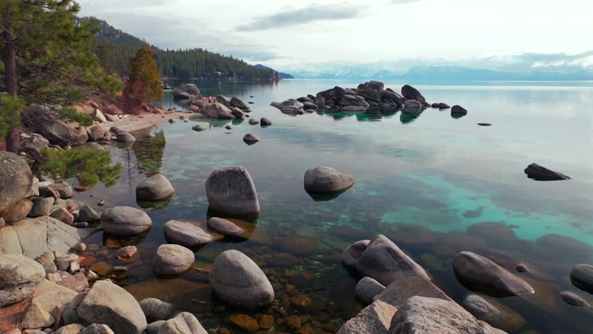 Lake Tahoe standing on rocky large boulders shoreline crystal clear glassy turquoise fresh water aerial drone Sand Harbor California sunny winter clouds Secret Chimney beach Incline Village static