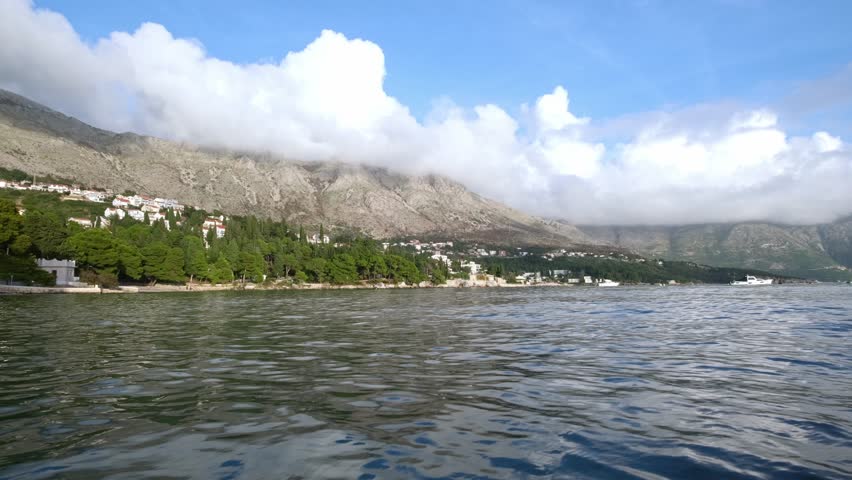 A static wide shot of a Croatia Coastline scene. Ocean in the foreground with trees on the farside shore. Then mountains behind and some clouds in the sky.