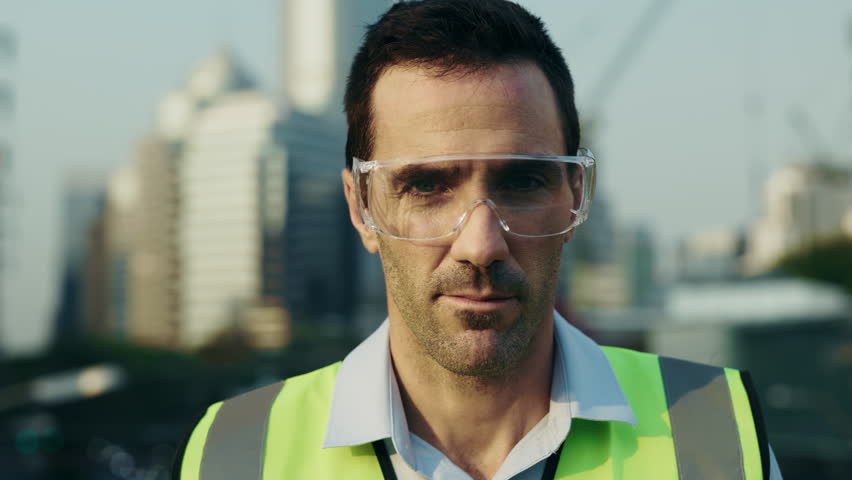 Portrait of man engineer in protective eyewear putting on safety helmet while looking at camera. Male builder in reflective vest adjusts hardhat preparing for construction tasks