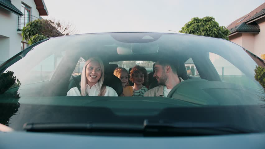 Woman in white shirt laughing while facing smiling man behind wheel. Two cheerful kids sitting close in backseat enjoying moment. Family bonding before heading out on relaxed neighborhood drive.