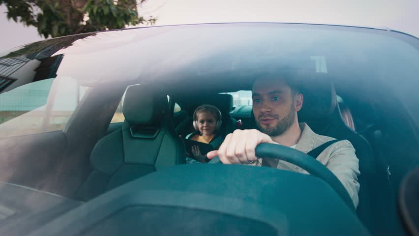 Smiling bearded male glancing back while driving and chatting with girl in car seat. Happy child wearing headphones holding tablet and reacting with excitement. Sharing fun moment during cozy ride.