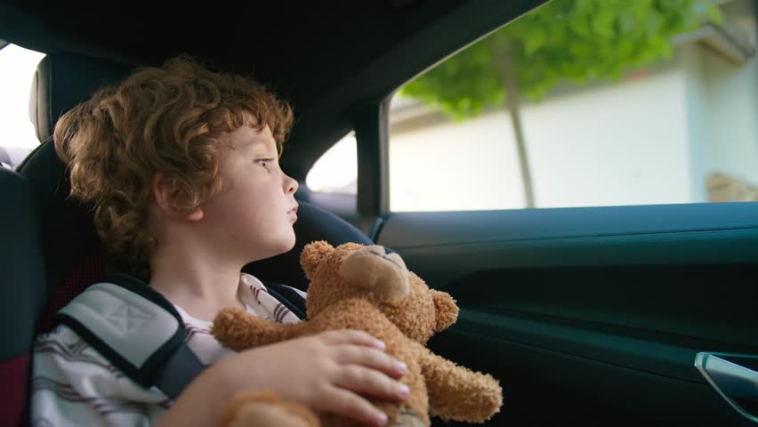 Curly-haired boy resting quietly in car seat holding brown teddy bear against chest. Gazing through window with calm. Enjoying peaceful drive past suburban homes in afternoon light.