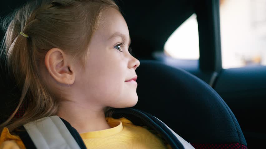 Adorable girl in yellow shirt smiling sweetly while sitting in child seat. Looking into camera with bright eyes and gentle expression. Enjoying safe cozy ride through town in back seat of car.
