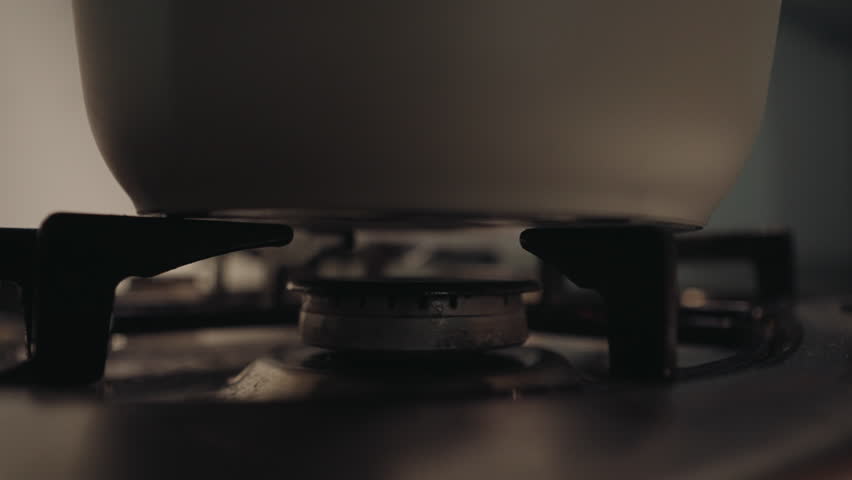 Close-up of unrecognizable person turning knob of gas stove in kitchen. Blue flame heating water in grey saucepan on gas stove, cooking and meal preparation in dark kitchen setting, slow motion.